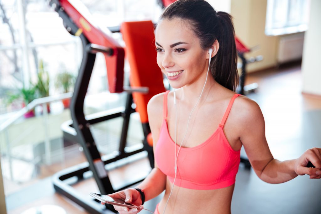 Cheerful attractive young woman athlete listening to music and using mobile phone in gym